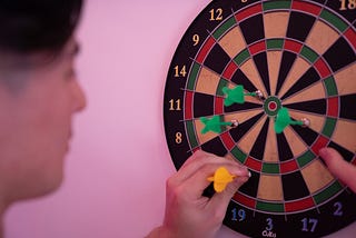 A group of darts clustered together on a dart board, with a man pulling out a yellow dart that wasn’t clustered together