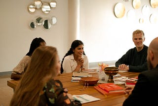 A group of five employees sit around a wooden meeting table talking, smiling, and laughing.