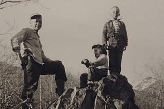 The author as a child sitting on a boulder eating an apple. Her brother and Dad stand above her. The family is on their homestead in Alaska.