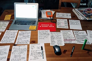 An open laptop sits on a wooden table covered with half sheets of handwritten notes