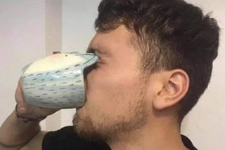 A person drinking from a blue mug with an unusual design, where the rim of the cup is curved inward, making it awkward to drink from. The mug’s shape blocks the person’s face, giving a humorous and impractical impression.