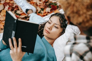 Hispanic woman lying on a blanket of red leaves reading a book.