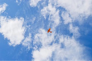 Small red plane against wide clouds in blue sky photographed from below