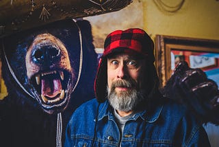 A bearded man with a funny facial expression while standing next to a growling stuffed bear