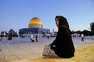 A woman wearing a black hijab sits on the ground with the dome of the rock in the background.