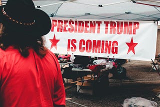 Person in cowboy hat and red shirt walking past a tent with a big sign reading “PRESIDENT TRUMP IS COMING”