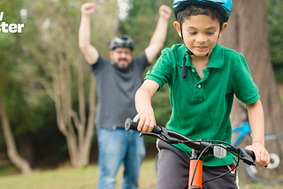 A child is riding a bike, an adult is celebrating behind him with his hands up in the air