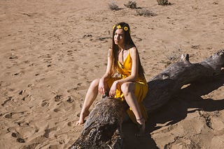 color photograph of a young woman wearing a yellow sundress sitting on a felled tree in the sand