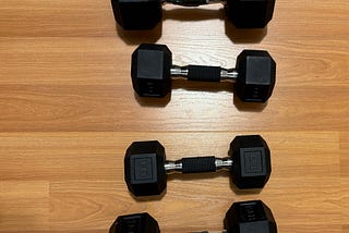 Four hexagonal dumbbells arranged in two columns on a wooden floor. The weights are labeled as 15 pounds and 10 pounds, with the 15’s  weight wrapped in tape on the handle for added grip. The dumbbells feature rubber-coated ends and metal handles with knurled grips.