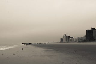 Moody photo of the ocean on the left & apartment buildings on the right, separated by a strip of sandy beach at Coney Island.