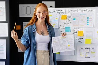 A photo of a young woman holding a page of diagrams and standing in front of a whiteboard covered with drawings and sticky notes.