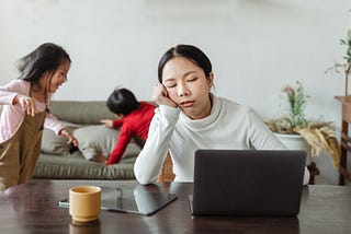 Young mom at a computer, while kids run rampant in the background. Mom’s eyes are closed, and she’s propping her head up with her arm leaning on the desk. She’s tired!