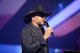 Jason Aldean speaking with attendees at the 2022 AmericaFest at the Phoenix Convention Center in Phoenix, Arizona on December 19, 2002. Aldean is wearing a black leather cowboy hat and a black leather jacket. He is holding a microphone, appearing to be speaking.