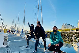 Image of three friends: Jessa takes a power stance with one arm open wide the other reaching down, Mariah crouches with her hands on her knees, and Geoff squats closer to the ground with his arms crossed and resting on his knees. The pose in front of their first keel boat.