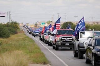 A convoy of pickup trucks flying “Trump 2020” flags stretches down a Texas road.