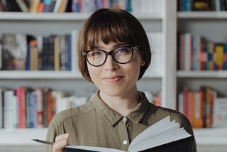 A woman with glasses looking up from a book with a pensive expression
