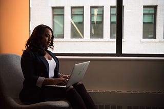 woman seated on a gray chair using a silver laptop in front of a window