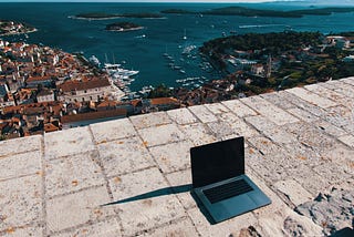 Laptop on brick wall with view of ocean and town behind it.