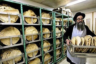 A bearded man wearing glasses and a black hat arranges matzahs on a four-layered shelf.