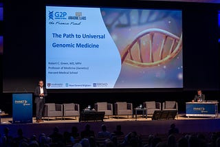 Dr. Robert C. Green stands at a podium in front of a screen displaying a title slide: “The Path to Universal Genomic Medicine.”