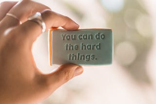 A left hand holding a small sign saying ‘You can do the hard things’