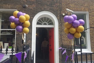 An open door in an arched doorway with a bundle of balloons floating on either side attached to the railings and bunting decorating the railings