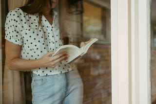 Linea reading a book through a window. She is wearing a white and black polka dot shirt.