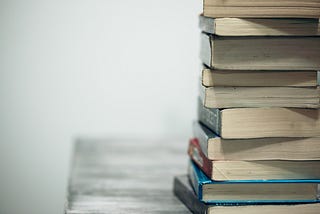 Books are stacked on a grey desk, taking up one side of the frame, their bottoms pointed toward the camera, no titles visible.
