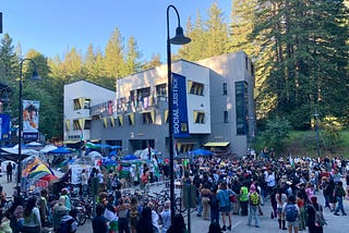 UCSC students gather at a rally for Rafah outside of the Palestine solidarity encampment on May 8. A university building, redwoods, and a blue sky are in the background. A banner on a lamp post in the foreground reads “Social Justice, The Real Change is Us.”