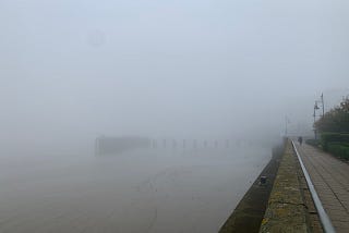 Heavy fog over the river, with a path stretching into the distance, and a jetty barely visible.