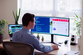 A professionally dressed man works at home with double computer screen and laptop on his desk with plants facing window