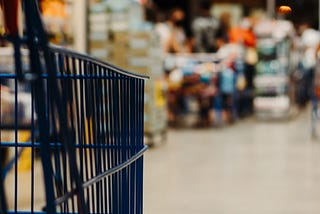 A close up picture of a blue shopping cart making it’s way to the check out aisle. The check out aisle in the background is blurred.