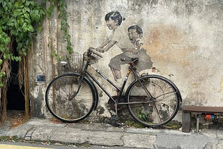 Penang street art — two kids on a bicycle — painted on a concrete wall with a real bicycle leaning against the wall.