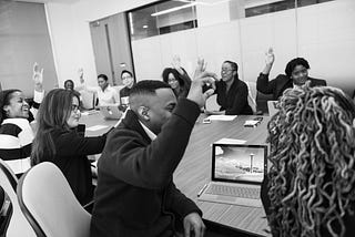 A black and white picture of several people sitting around a conference table, many of them have their hands raised. They are a variety of genders and races.
