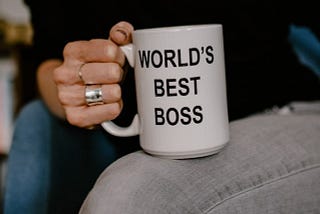 Hand of a woman in black holding white ceramic mug with the text “World’s Best Boss”.