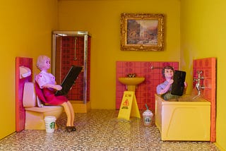 Two dolls in a tiny pink and yellow bathroom, one on the toilet and one in the bathtub, both looking at giant smartphones.