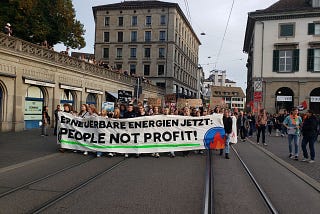 Ein Fronttransparent — Erneuerbare Energien Jetzt! People not Profit mit dem Klimastreiksymbol. Demozug dahinter auf einer Strasse in Zürich