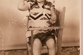 Old black & white photo of little boy sitting in a chair and holding a ball.