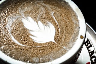 Close up of a flat white coffee with latte art in the shape of a feather, on a “Black and White” branded cup and plate.