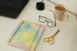 Photo of a computer keyboard and mouse, spiral bound journal, scissors, glasses, cup of coffee.