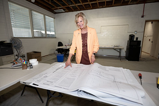 In a room full of tables, boxes, and fans, a woman is in the center of the photo and she is pointing at a long white table. The table has several large papers and appears to be instructions for a possible layout. The slightly older woman is smiling and is wearing a yellow jacket with a brown shirt.