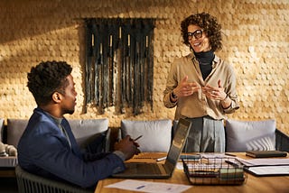 A man sitting at a wooden table working on his laptop looking up to a woman who is standing beside the desk speaking with him. They are in a yellow sunny room with a grey couch.
