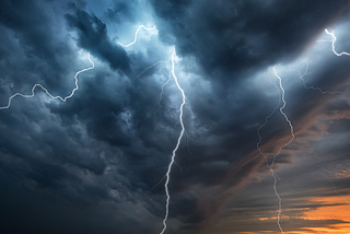 Lightning bolts in dark storm clouds against an orange sunset.