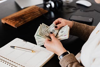 Hands counting a stack of money with a notebook and pen on the table.