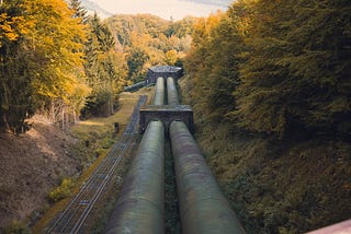 a pipeline running alongside a single-track train track. On other side of the pipeline and the train track is a thick forest. In the distance, there are forest-covered mountains