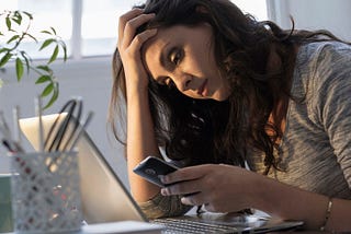 A millennial woman who feels depressed sits at her desk, clutching her forehead and looking at her phone.