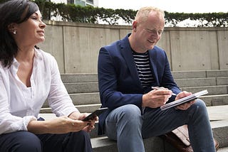 a man in a blazer sits next to a woman in a white shirt smiling at his notepad after learning how to balance his heart-centered work and marketing practice