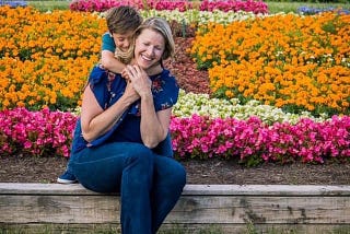 Single Mother by Choice- woman with toddler son hugging her from behind as she sits on the edge of a flowerbed with pink, yellow and orange flowers blooming