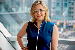 Sarah Kurchak, a white woman with blonde hair and white glasses, smiles into the camera for a portrait. She wears a dark blue shoulderless zip-up top and stands in front of a window that looks out over a city junction.