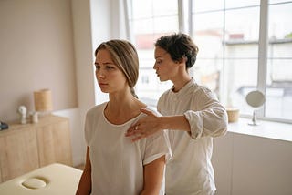 A therapist gently evaluates a seated woman’s shoulder posture during a physical therapy or body alignment session. The setting is calm and bright, with natural light streaming through large windows. Reflecting the perspective of Ramneek Bhogal on the holistic approach to managing tennis elbow.